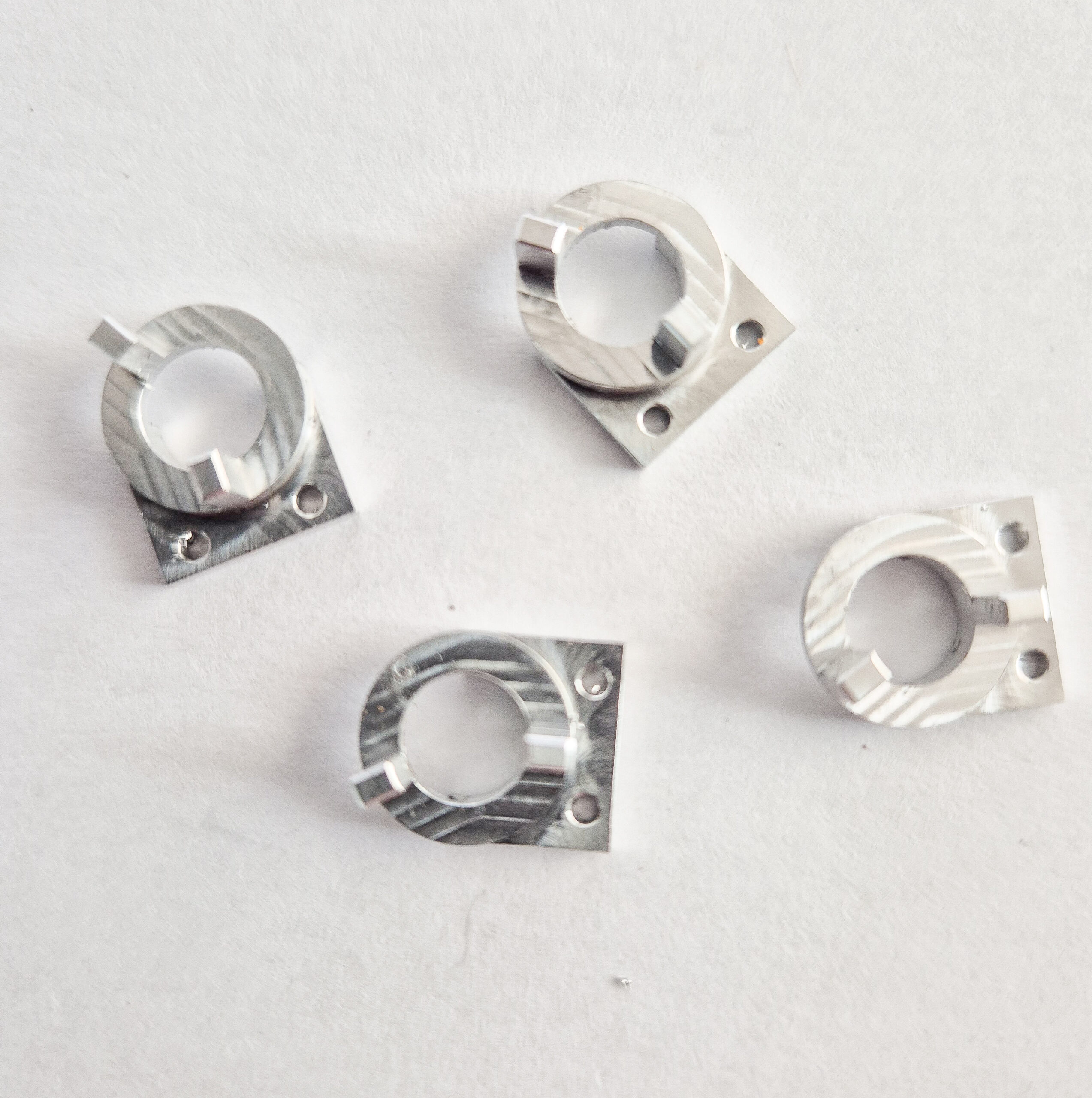 Quatre petites pièces métalliques usinées en aluminium, de forme semi-circulaire avec des trous de fixation